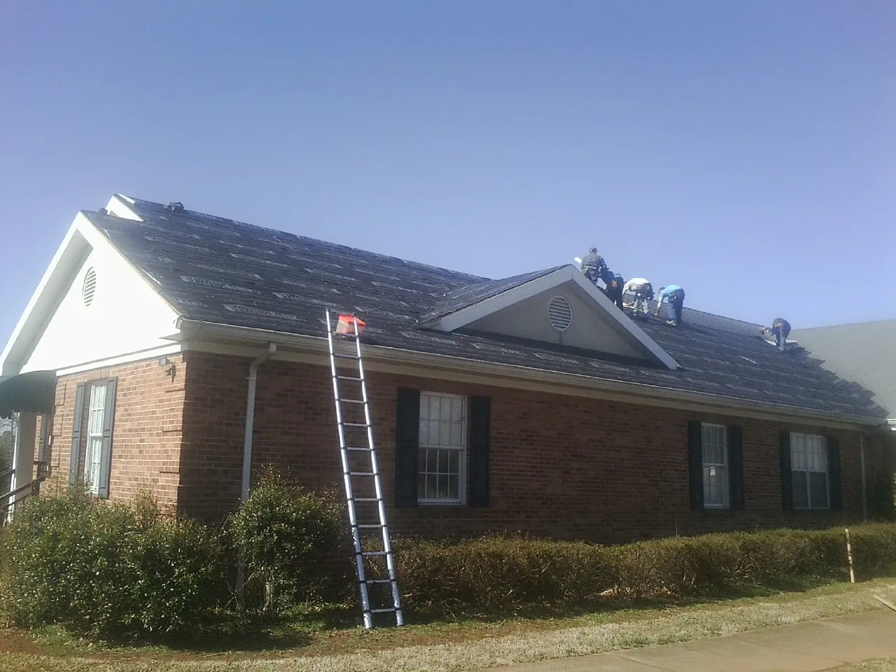 Crew tearing shingles off a brick ranch roof with a ladder set — Emergency Roof Repair work by our crew in Bergheim
