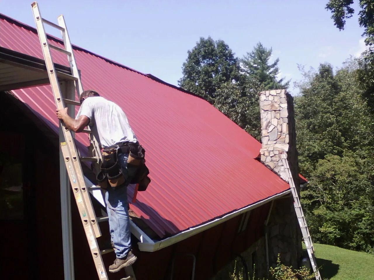 Roofer on a ladder beside a red metal roof and stone chimney — Soffit & Fascia Repair by Roofing Brothers in Bergheim